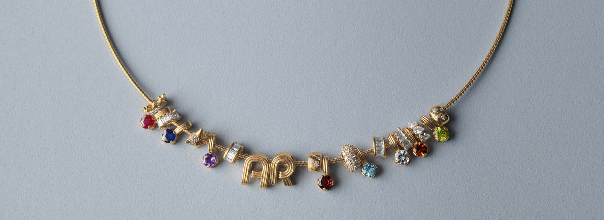 A gold chain necklace with a central cluster of small gold charms and colorful gemstones, laid flat on a light gray background.