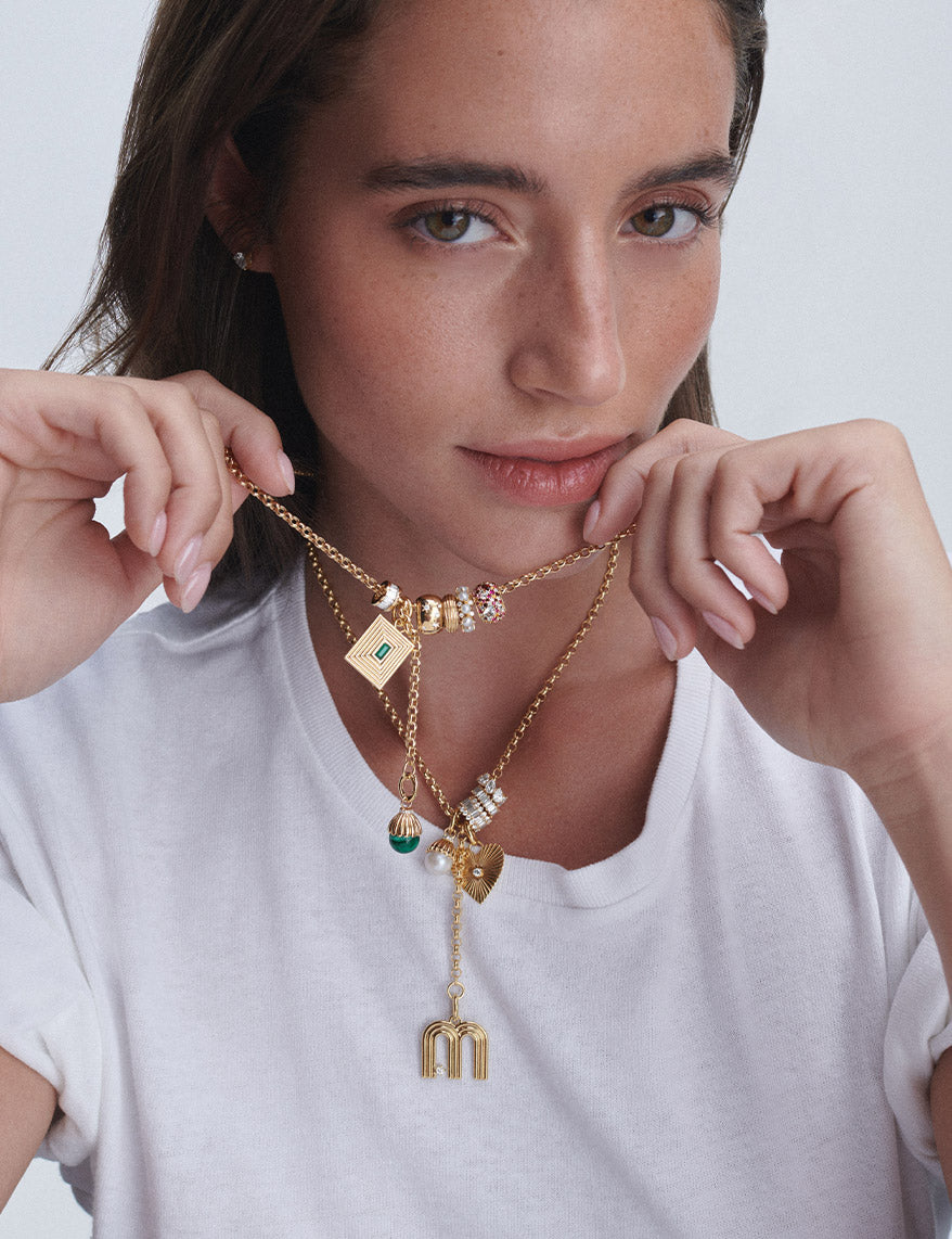 A close-up of a woman wearing a layered gold necklace set with various charms, including a square pendant with a green diamond, a heart, and a small hanging letter 'M' charm, against a light gray background.