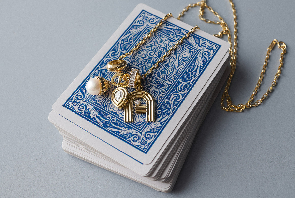 A gold-toned necklace with a heart-shaped pendant, a small white pearl bead, and a circular charm, draped over a stack of blue-backed playing cards on a light gray surface.