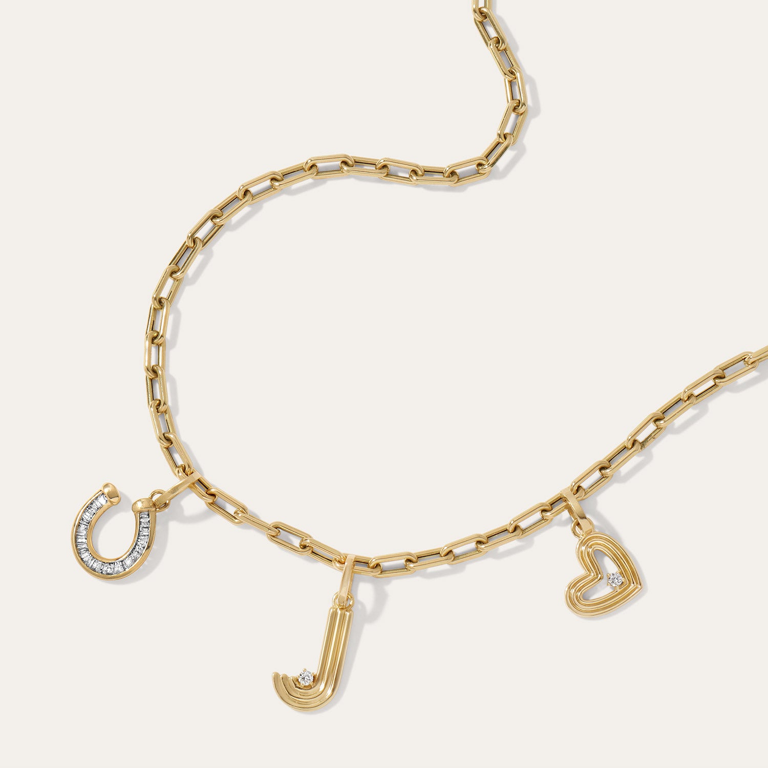 A gold chain necklace with three charms: a horseshoe, a letter 'J', and an open heart, each accented with one small diamond, displayed on a light cream background.