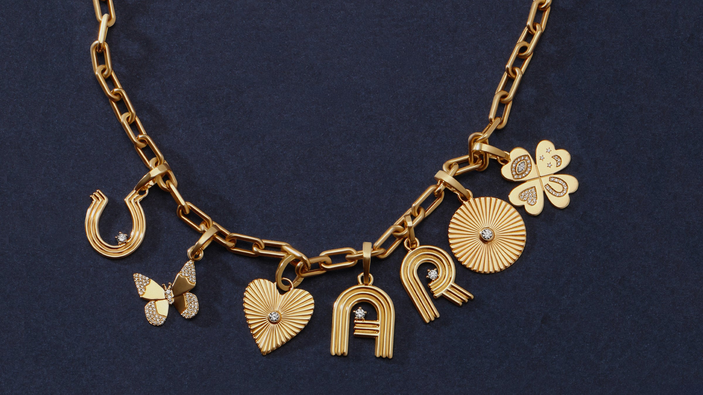 A gold chain necklace with multiple charms, including a horseshoe, butterfly, heart, sunburst, clover, and letters in 'A' and 'R', displayed on a dark navy blue textured background.
