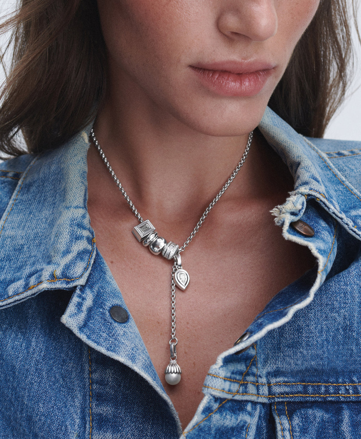 A close-up of a model wearing a silver lariat-style necklace with charms, including a small heart pendant and a dangling bead, paired with a blue denim jacket.