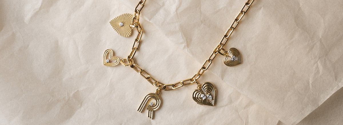 A gold-tone chain necklace with multiple heart charms and a letter 'R' charm, laid flat on a textured beige paper background.