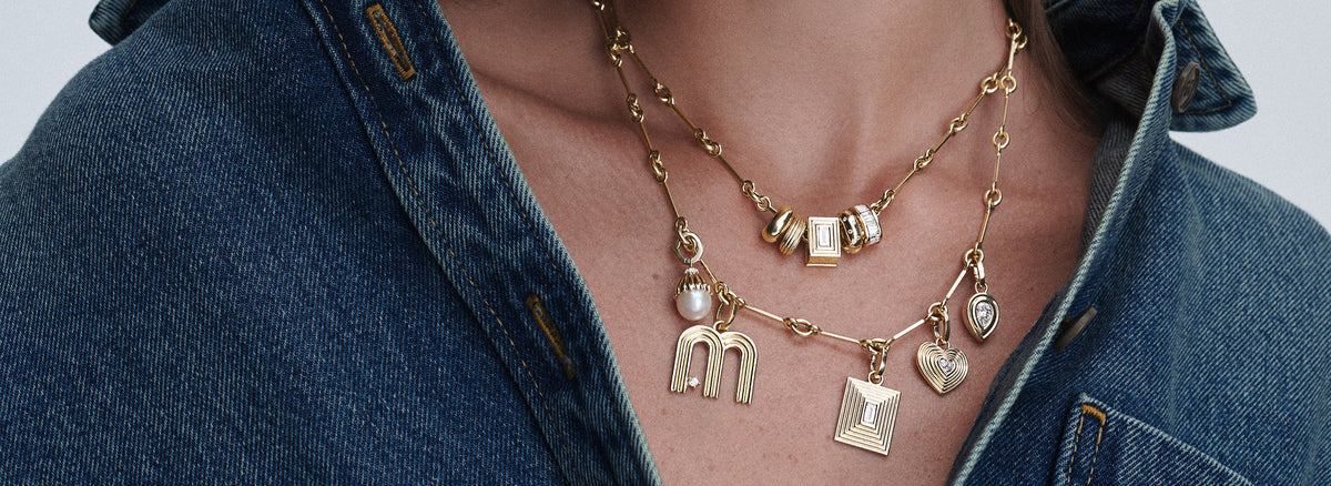 A close-up of a person wearing a layered gold necklace with multiple charms, including letters and geometric shapes, styled with a blue denim shirt.