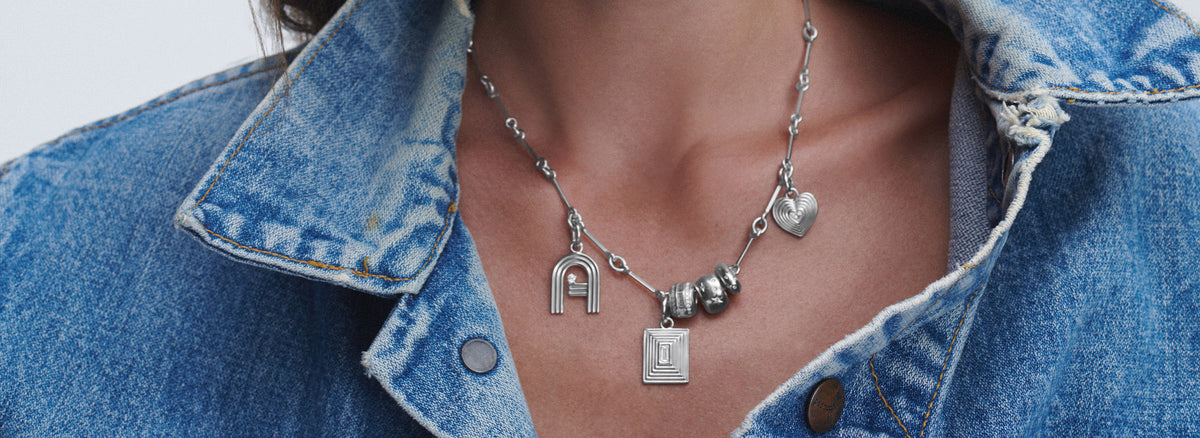A close-up of a person wearing a silver charm necklace with a heart, letter 'A' charm, styled with a blue denim jacket.