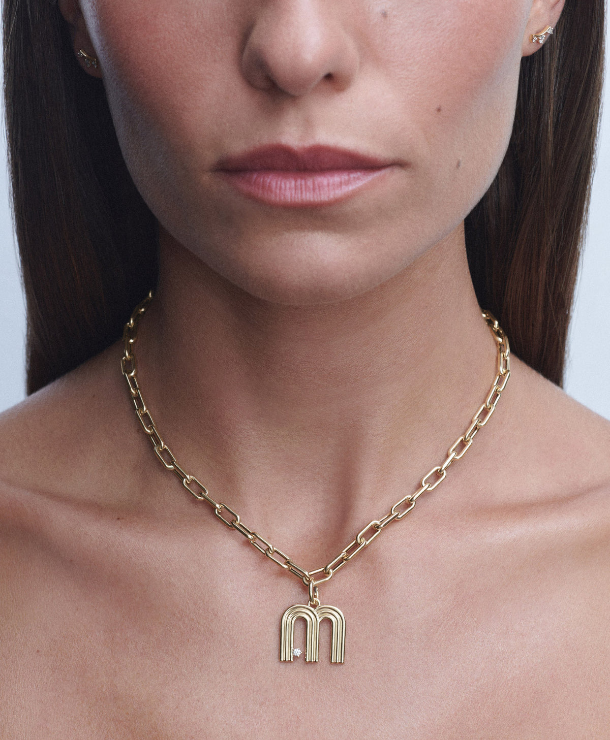 A close-up of a woman wearing a gold chain necklace with a stylized 'm' pendant, shown against her neck and shoulders.