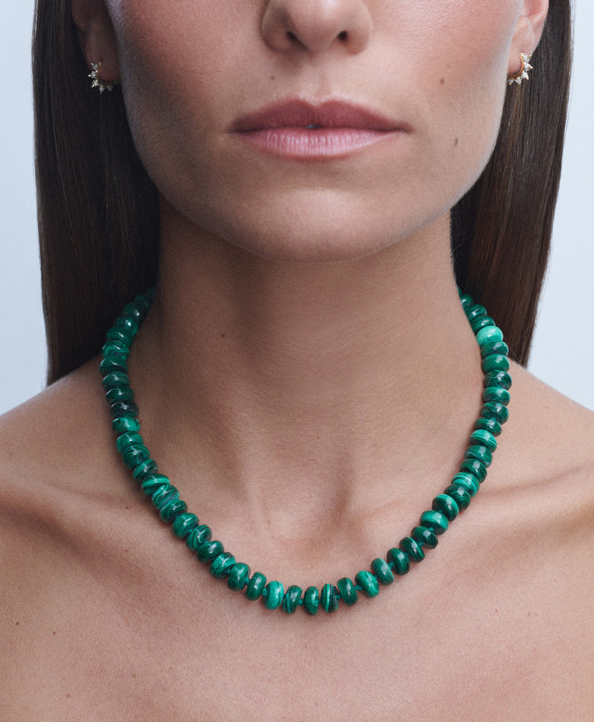 A close-up of a model wearing a green beaded necklace, paired with small gold stud earrings.