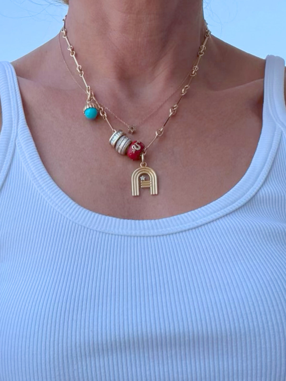 A close-up of a person wearing a layered gold necklace with a letter 'A' pendant and colorful beads, paired with a light blue ribbed tank top.