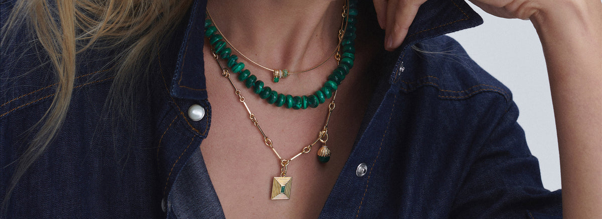A close-up of a person wearing layered necklaces, including a green beaded choker, a gold chain with a green bead, and a gold chain with a square pendant, styled with a dark denim shirt.