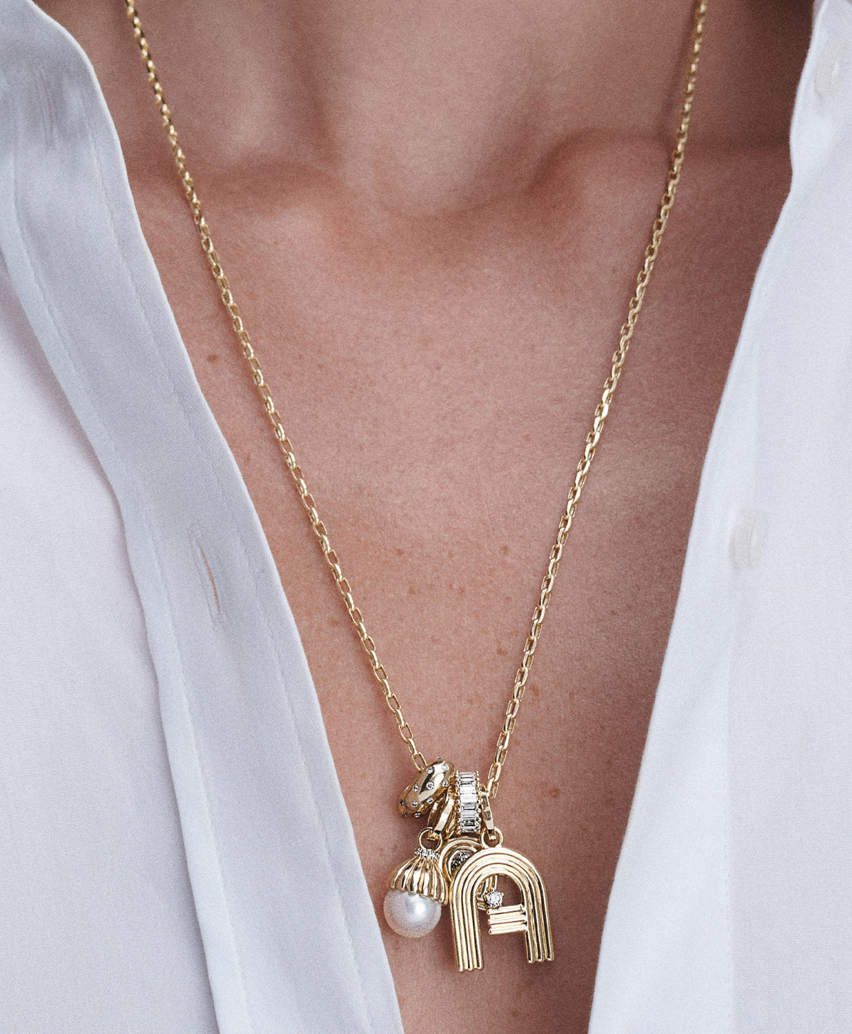 A close-up of a gold chain necklace with a multi-charm pendant worn by a model. The pendants features a gold letter 'A' charm, a pearl drop charm, and two diamond big beads.
