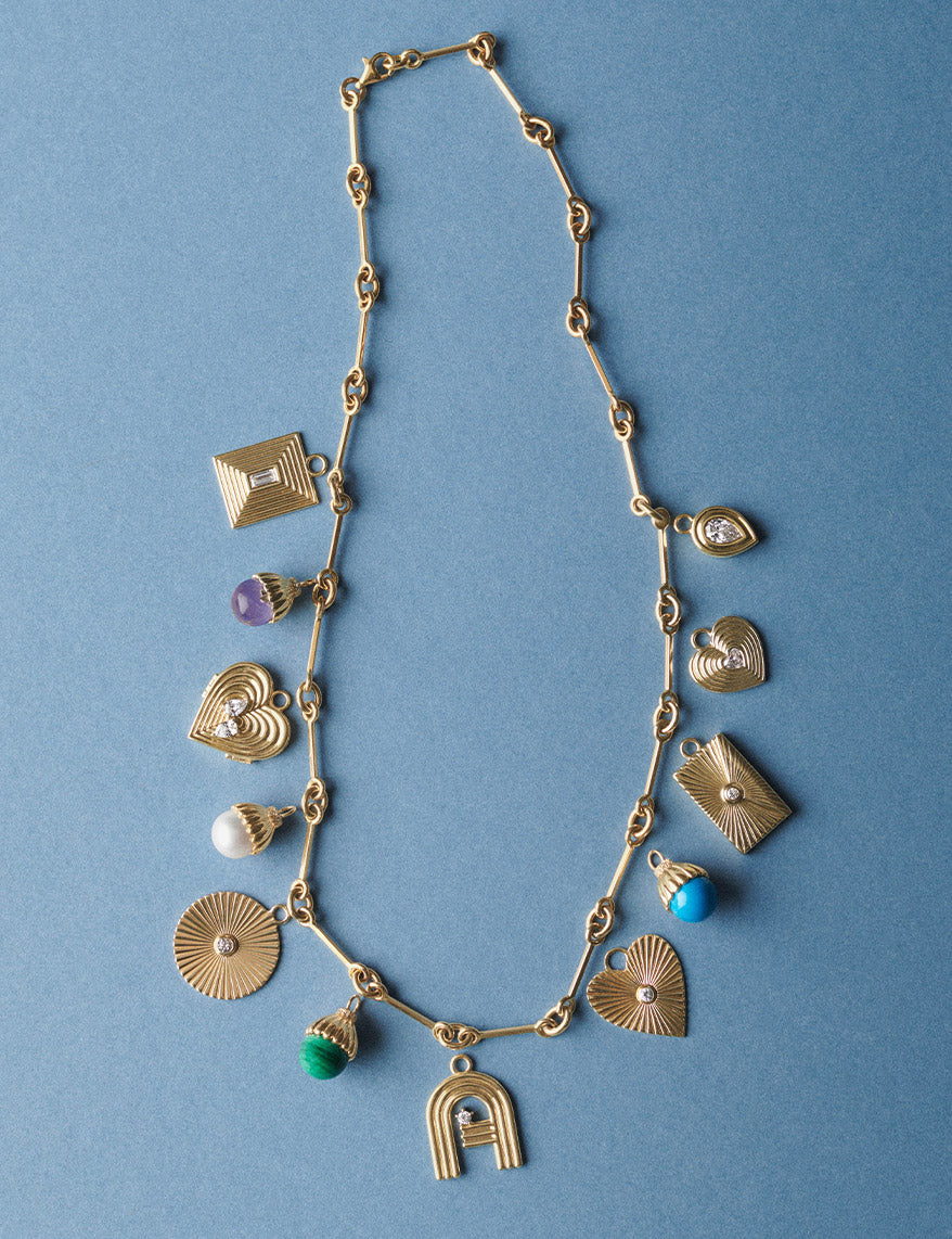 A gold-toned link chain necklace with multiple dangling charms, including heart, sunburst, shell, and abstract shapes, accented with small colorful stones, photographed on a solid blue background.
