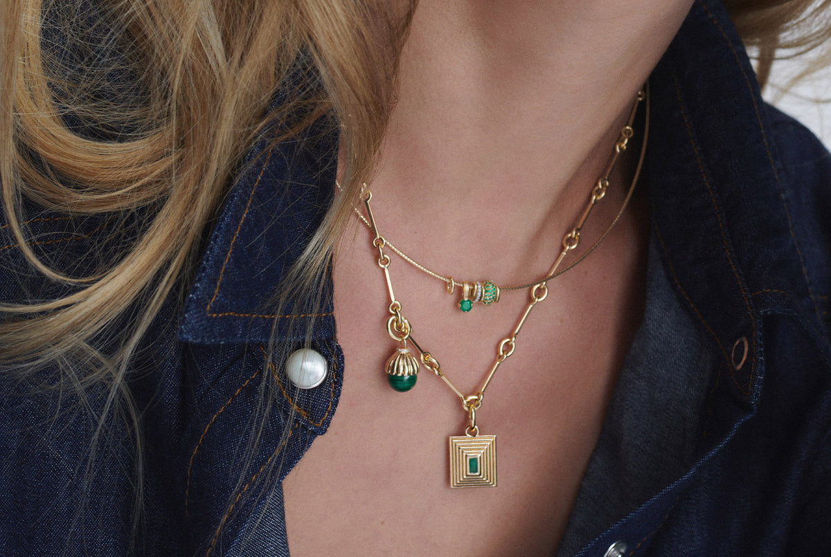 A close-up of a model wearing a layered gold necklace set with small green stone accents and a square pendant, styled with a dark denim shirt.