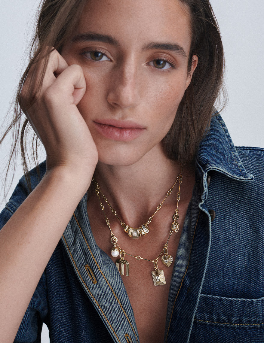 A close-up of a woman wearing layered gold necklaces with various charms, styled with a denim shirt.