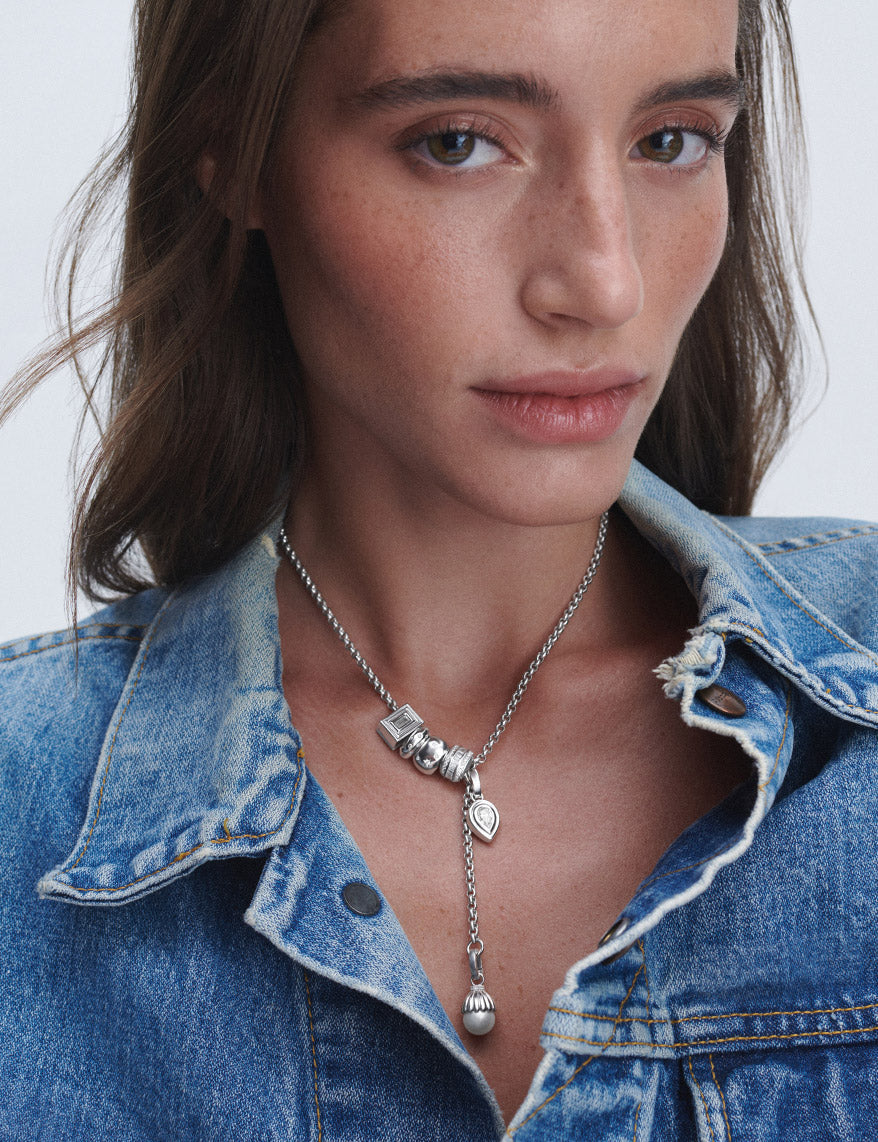 A close-up of a woman wearing a silver charm necklace with multiple big beads and charms with a pearl and diamonds, styled with a blue denim jacket.