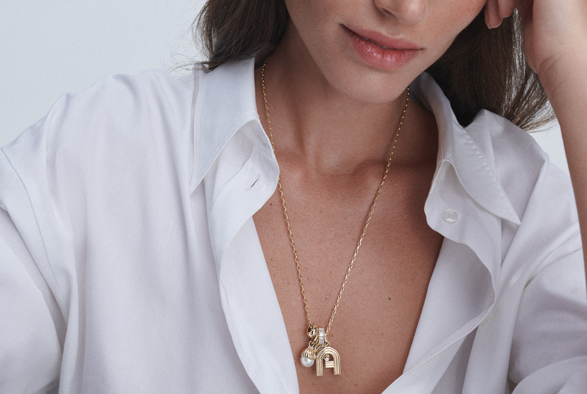 A close-up of a woman wearing a delicate gold chain necklace with a small gold initial 'A' pendant and a pearl charm, styled with a white button-down shirt.