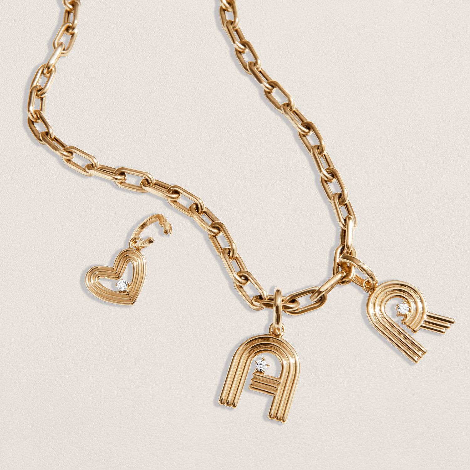 Gold-tone chain necklace with three charms: a heart-shaped charm with a diamond, and two stylized letter 'A' and letter 'R' pendants, both with diamonds, displayed on a light beige background.