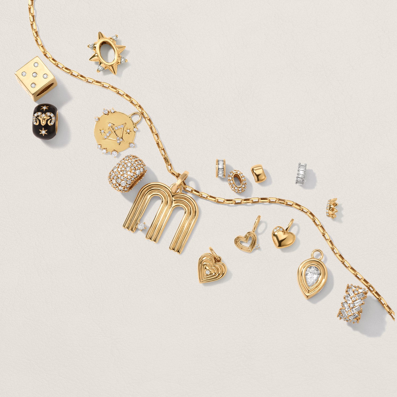 A gold chain necklace with multiple gold, silver, and diamond charms, including hearts, stars, a dice,a crown, letters, and a spike, arranged on a light beige background.