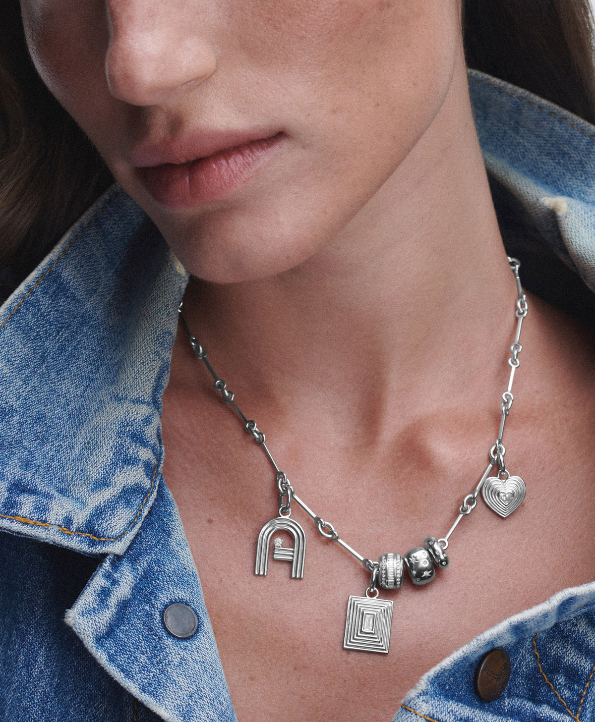 A close-up of a model wearing a silver chain necklace with multiple charms, including a heart, a square with a letter 'D', and a letter 'A' charm, styled with a denim jacket.