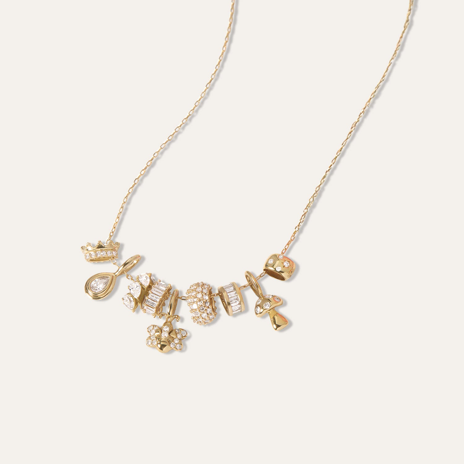 A delicate gold chain necklace featuring a cluster of small, whimsical gold and diamond charms, including a teardrop, a crown, and a flower, displayed on a clean off-white background.