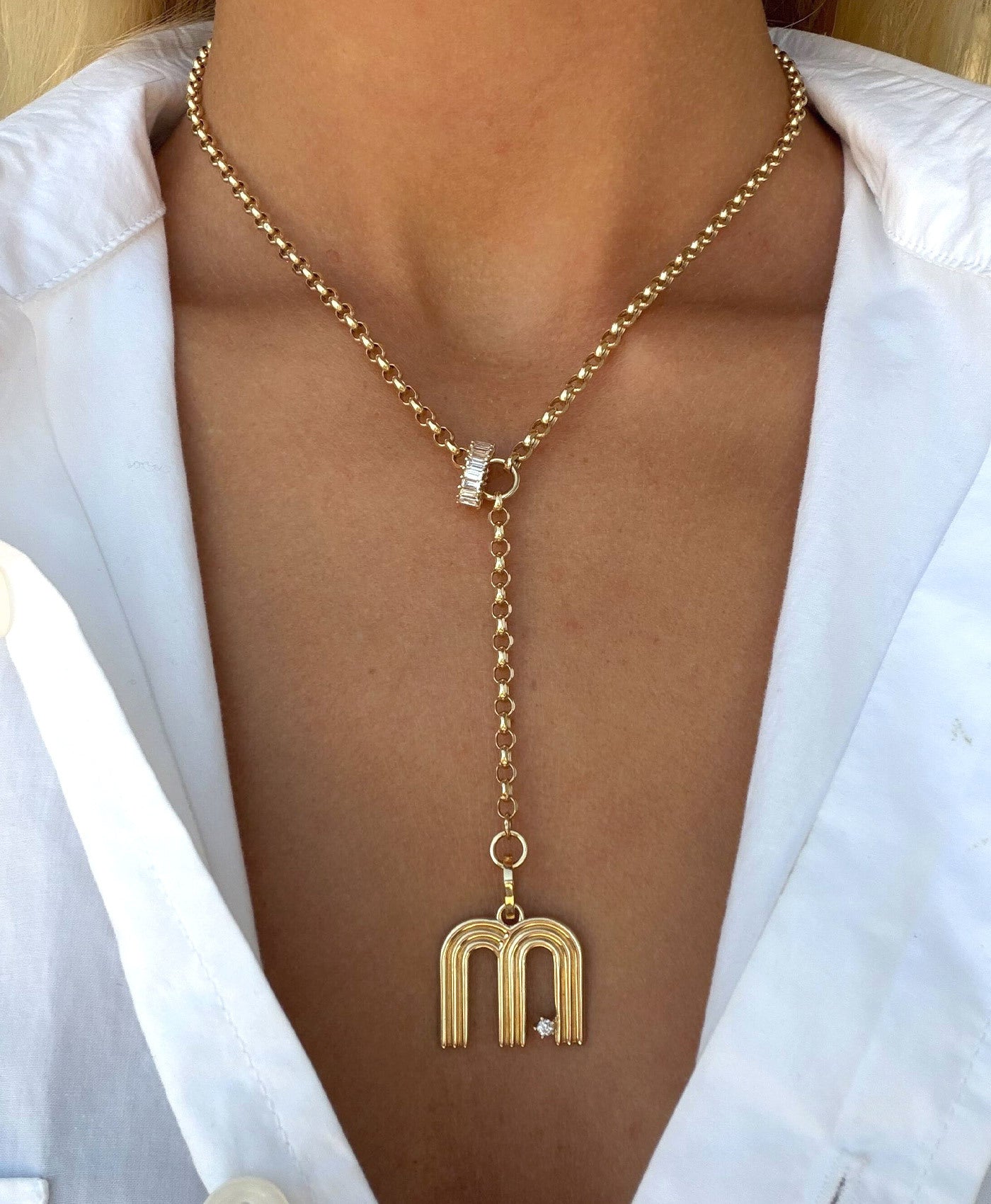 A gold lariat-style necklace with a letter 'M' charm and a diamond big bead pendant, shown worn on a model's neck over a white collared shirt.