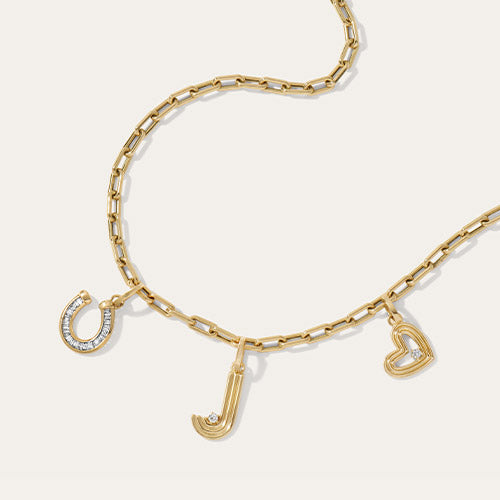 A gold chain necklace with three charms: a horseshoe, a letter 'J', and an open heart, each accented with one small diamond, displayed on a light cream background.