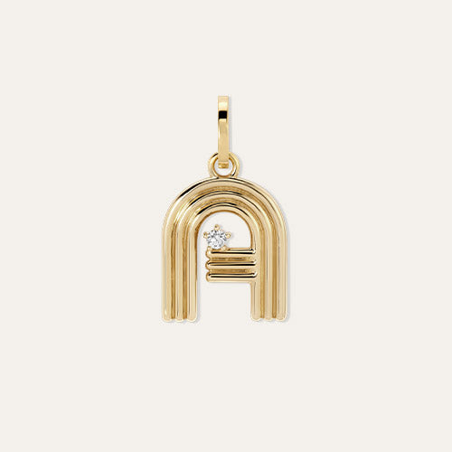 A gold charm featuring a letter 'A' design with a small diamond accent, shown on a light cream background.