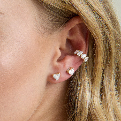A woman with blonde hair with two diamond heart stud earrings and one diamond ear cuff on ear.