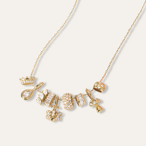 A delicate gold chain necklace featuring a cluster of small, whimsical gold and diamond charms, including a teardrop, a crown, and a flower, displayed on a clean off-white background.