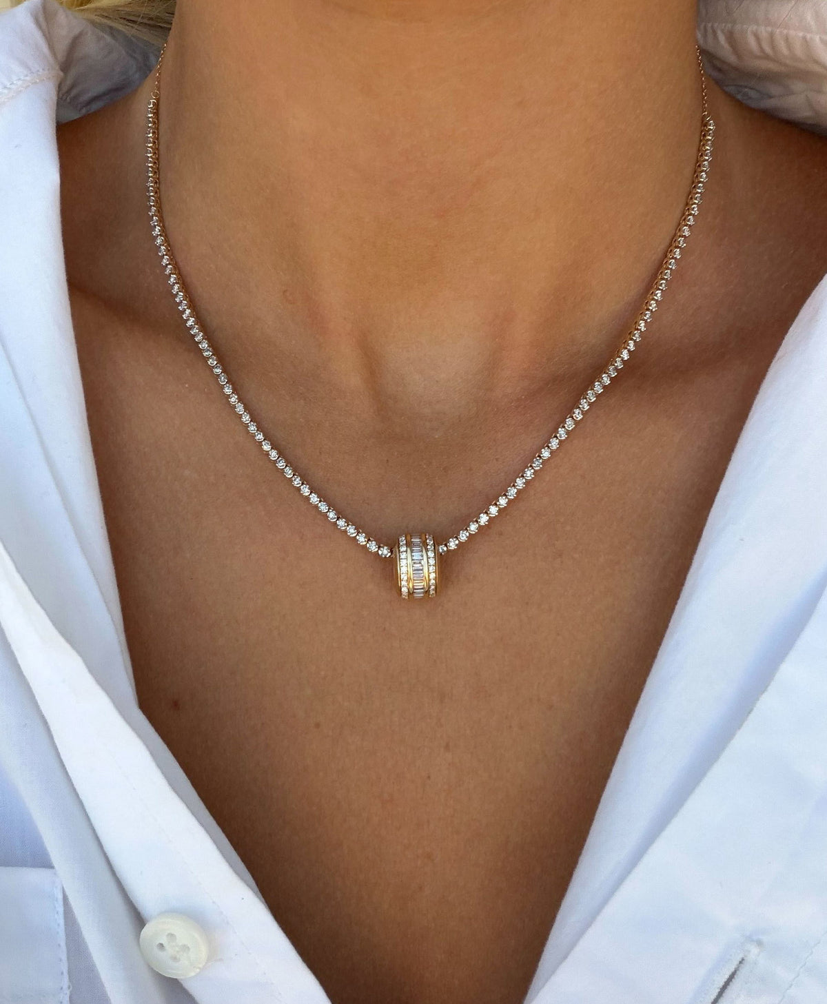 A close-up of a diamond tennis necklace with a gold and diamond big bead pendant, worn by a model in a white collared shirt.
