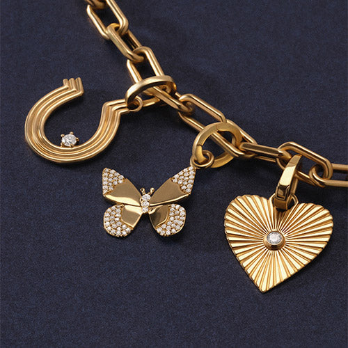 A gold chain necklace with multiple charms, including a horseshoe, butterfly, heart, sunburst, clover, and letters in 'A' and 'R', displayed on a dark navy blue textured background.