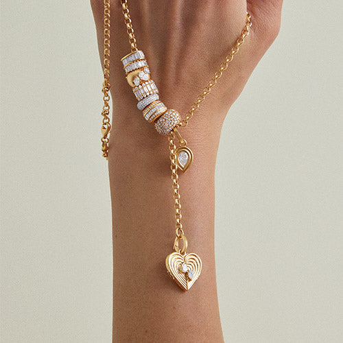 A close-up of a gold Y-necklace with a heart-shaped pendant and diamond big beads, worn on a model's wrist against a neutral beige background.