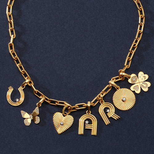 A gold chain necklace with multiple charms, including a horseshoe, butterfly, heart, sunburst, clover, and letters in 'A' and 'R', displayed on a dark navy blue textured background.