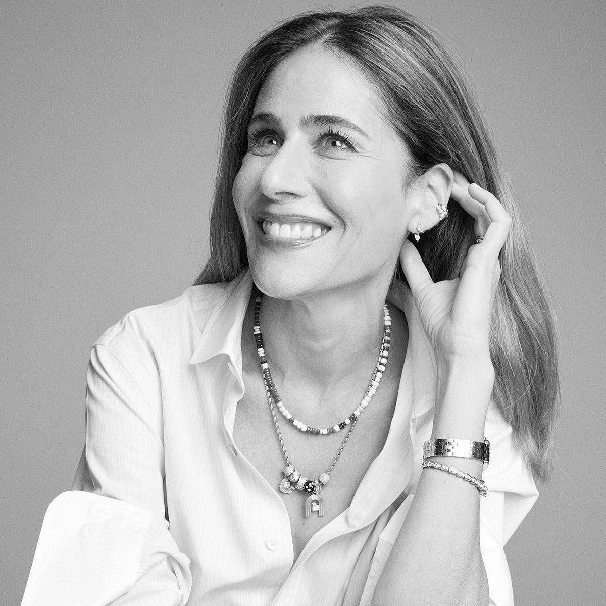 A black and white portrait of a smiling woman wearing layered necklaces, bracelets, and earrings, wearing a white button-down shirt against a neutral gray background.
