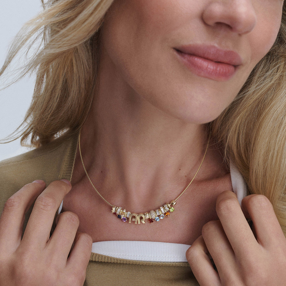A close-up of a person wearing a gold necklace with colorful gemstone accents and a central letter charm, styled over a white shirt and tan cardigan.
