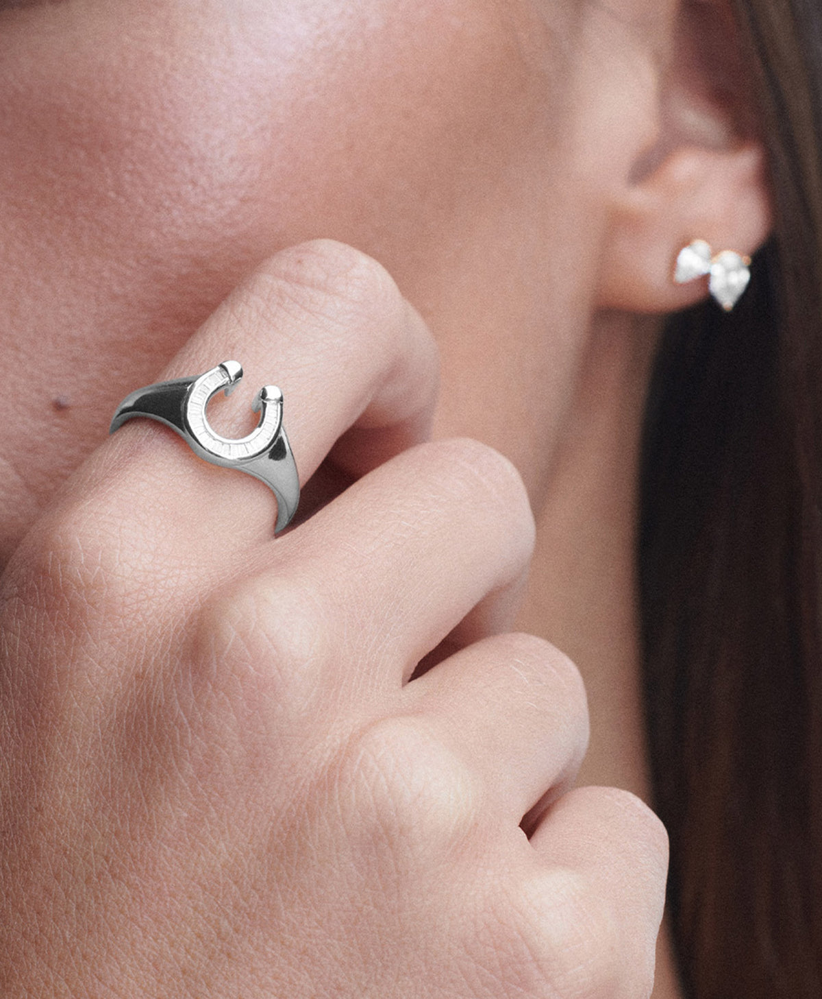 A close-up lifestyle shot of a model wearing a silver horseshoe ring on her finger, with a silver stud earring with diamonds visible on her ear.